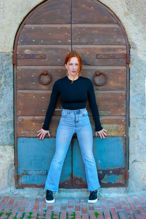 Free A stylish redhead woman poses confidently against an aged wooden door, showcasing a blend of modern fashion and rustic charm. Stock Photo
