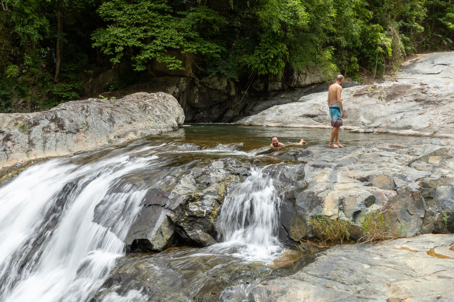 Two men enjoying a natural waterfall pool surrounded by lush greenery and rocky formations.