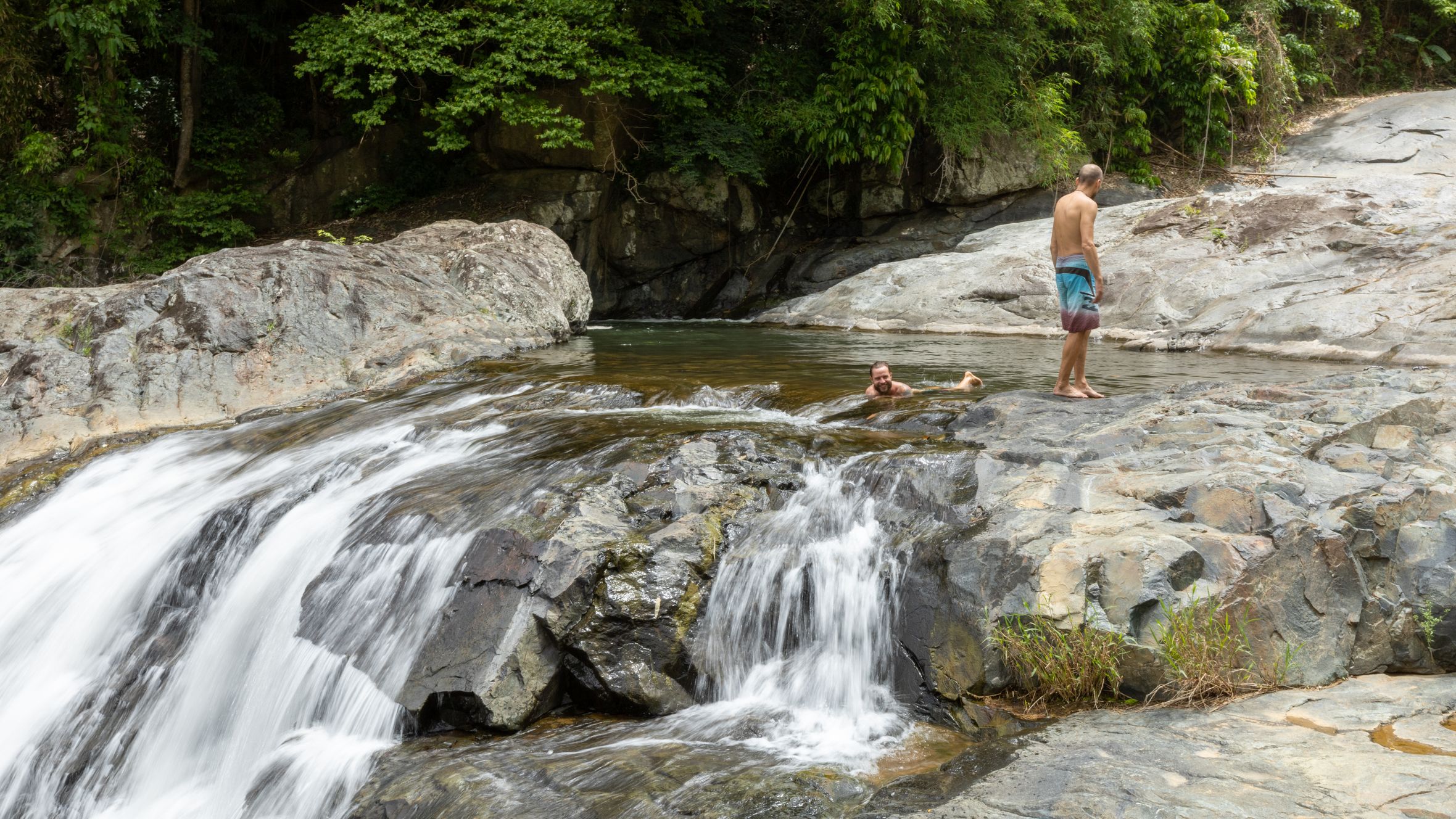 Two men enjoying a natural waterfall pool surrounded by lush greenery and rocky formations.