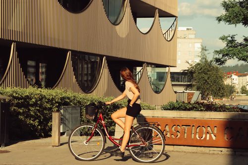 Free Woman cycling past modern building in sunlit Jönköping, Sweden. Urban lifestyle captured in vibrant scene. Stock Photo