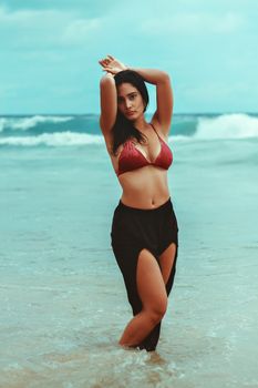 Elegant woman posing with arms raised on a serene beach, evoking fashion and vacation vibes.