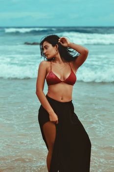 A beautiful woman in a bikini and skirt posing gracefully on a sandy beach with waves.