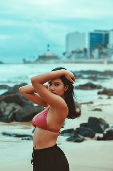 Portrait of a woman with long hair posing confidently by the rocky shore, against an urban backdrop.