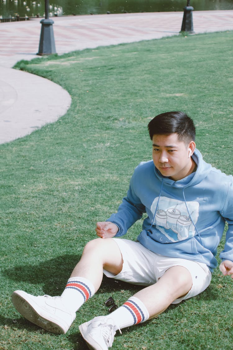 Young Man In Blue Hooded Sweatshirt And White Shorts Sitting On Lawn