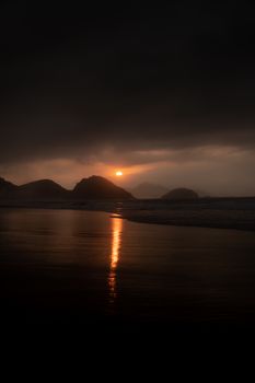 A dramatic sunset with reflections over a calm beach, creating a silhouette of distant hills.