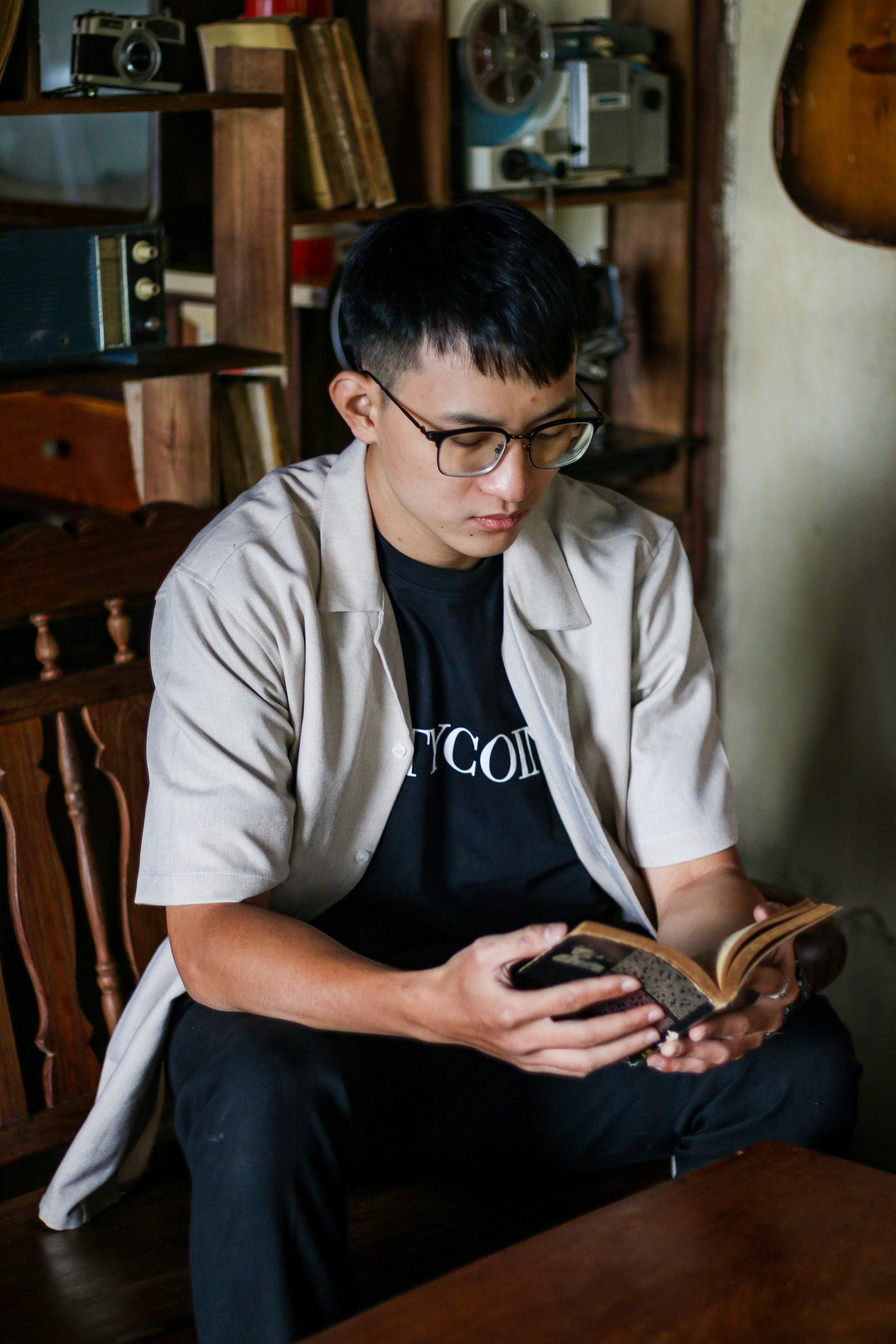 Man in glasses reading a book indoors, surrounded by vintage decor, creating a calm and introspective setting.