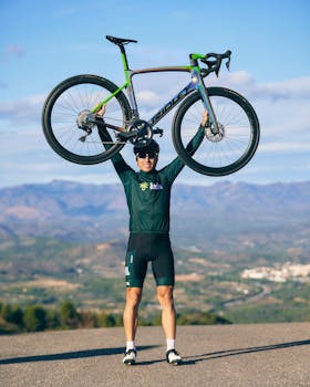 Man in cycling gear lifts road bike victoriously outdoors with scenic mountains in background.
