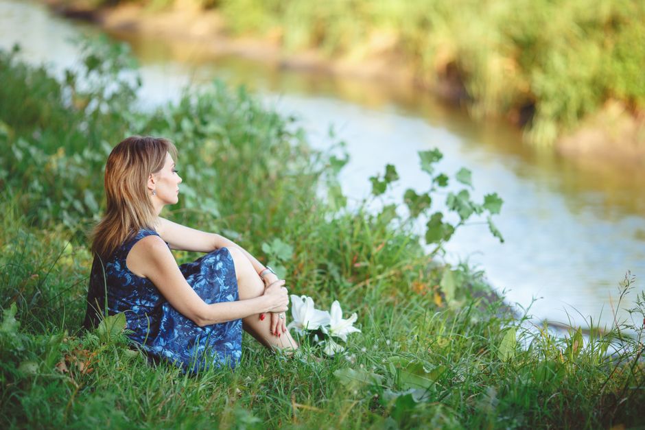 A woman in a blue dress sits peacefully by a scenic riverside, embracing the tranquil outdoor setting.