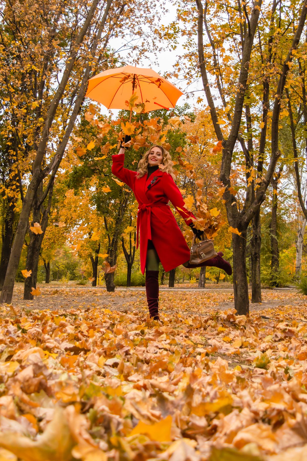Woman in red coat and umbrella posing amid vibrant autumn leaves in a park.