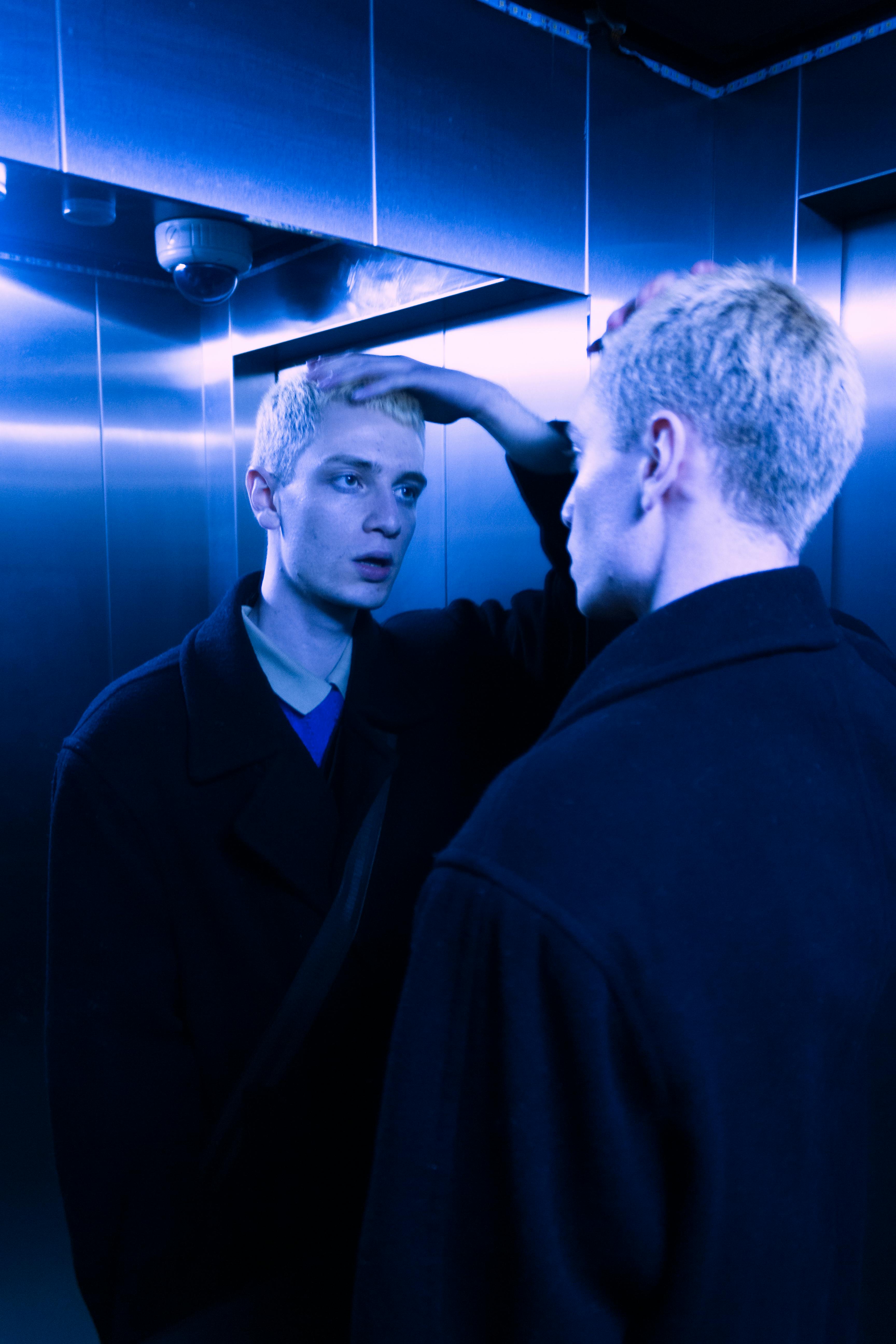 Man with blonde hair reflecting in elevator mirror under blue light.