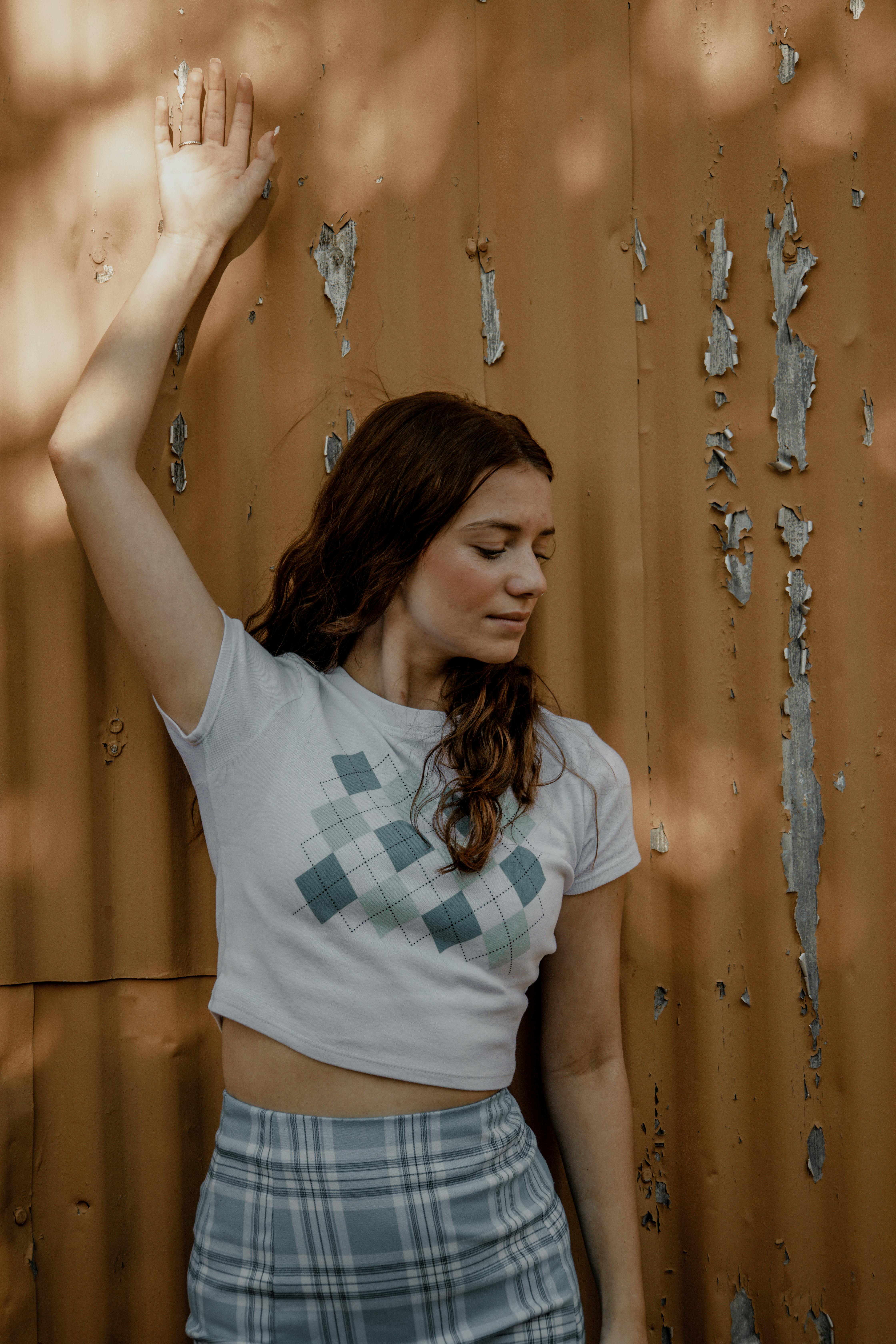 Woman in a plaid skirt and graphic tee standing against an aged wall, enjoying the sunlight.