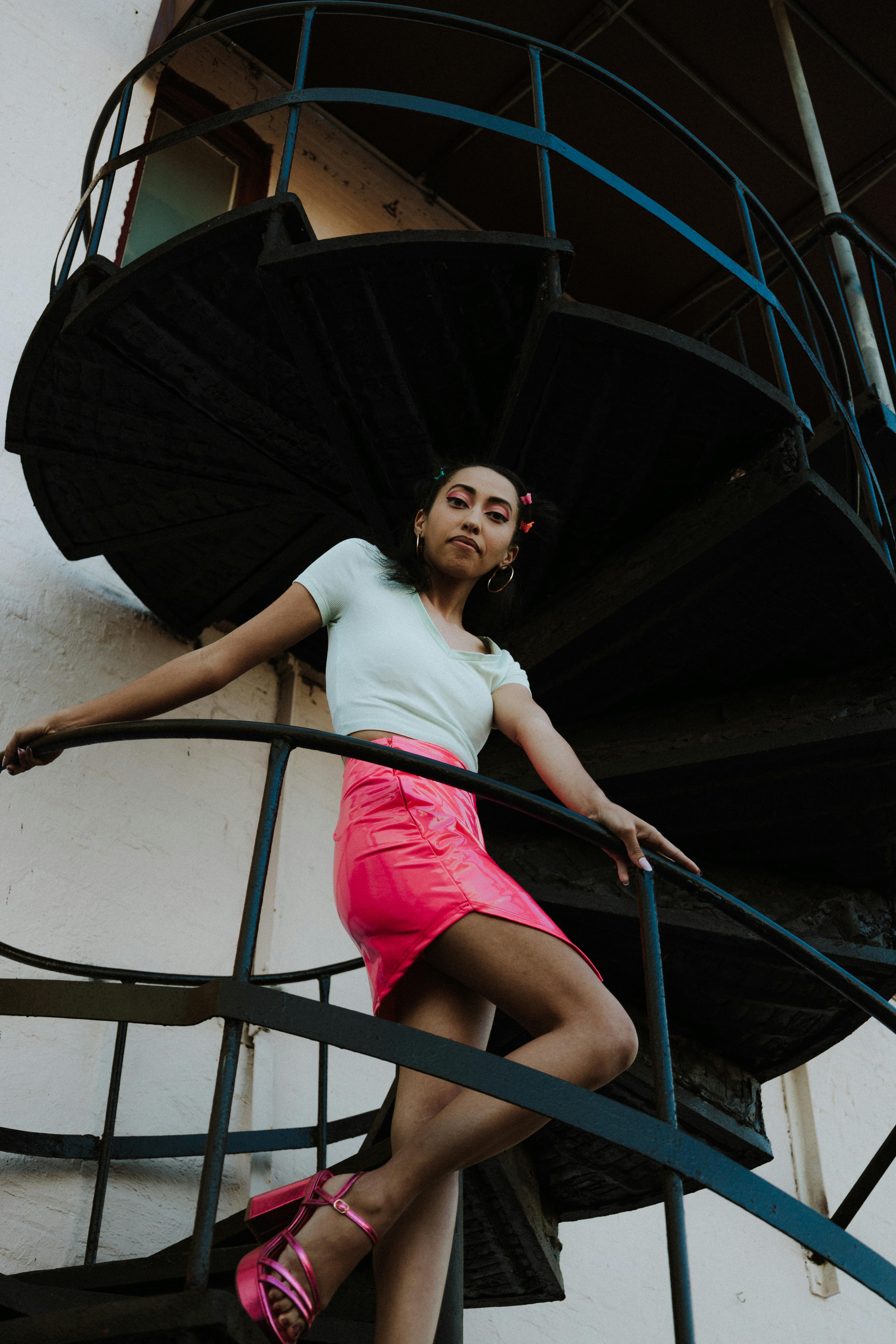 a woman in a pink skirt and white top is standing on a spiral staircase