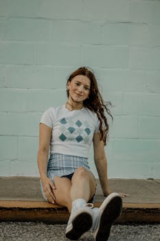 A young woman sitting casually outdoors against a pastel wall. Stylish and relaxed atmosphere.