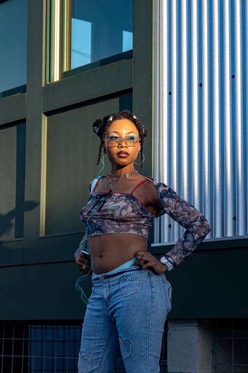 Free Fashionable young woman poses confidently outdoors during golden hour, wearing trendy sunglasses and a unique outfit. Stock Photo