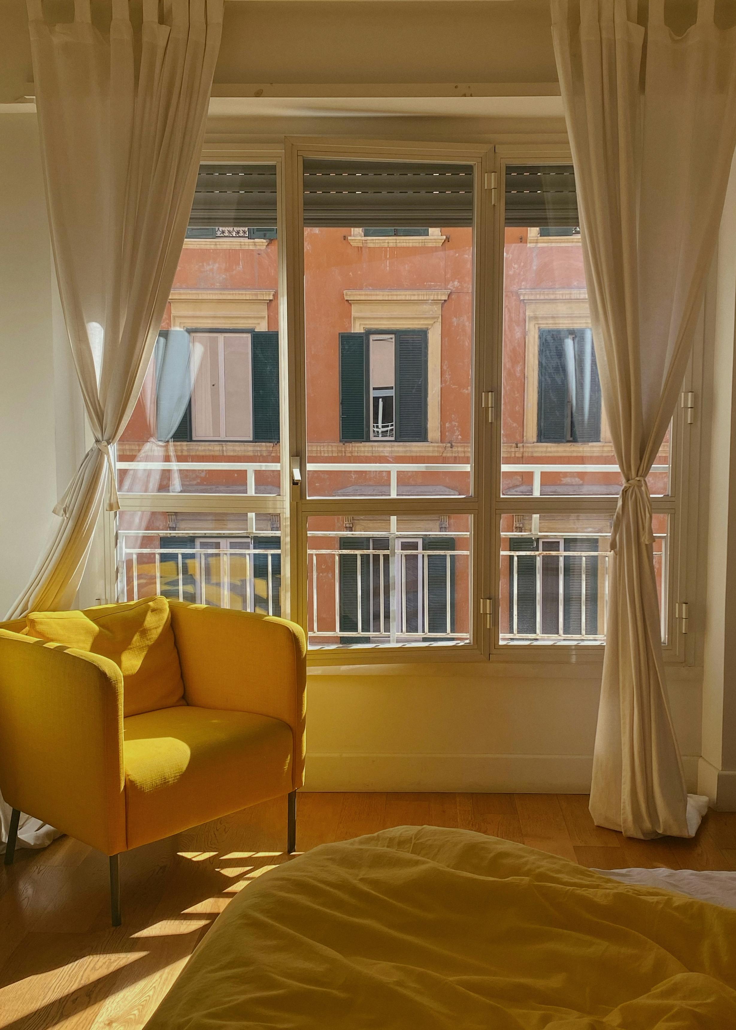A bright interior featuring a comfortable yellow armchair, large windows, and white curtains.