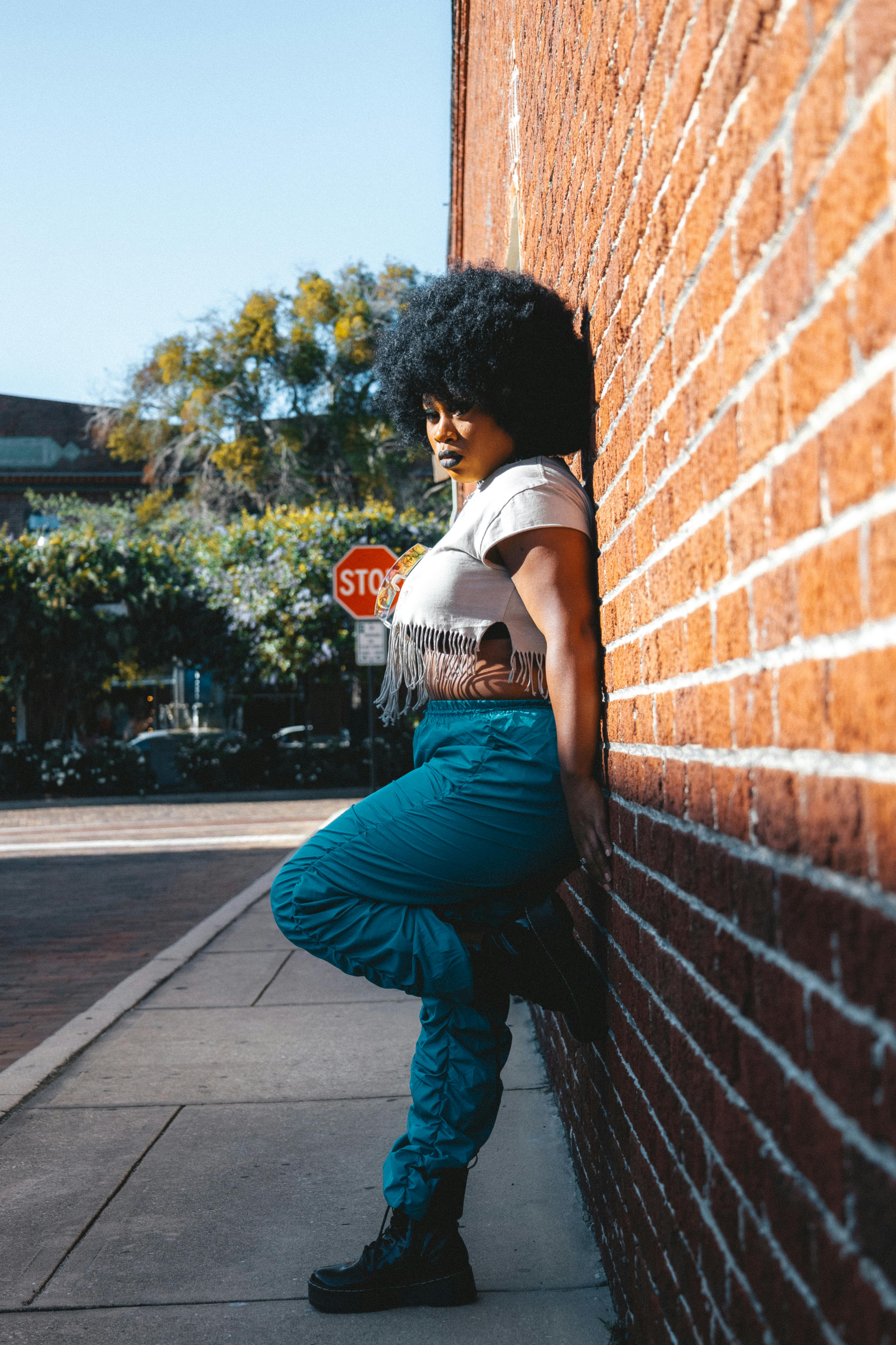 A fashionable woman leans on a brick wall under bright sunlight, capturing urban street style.