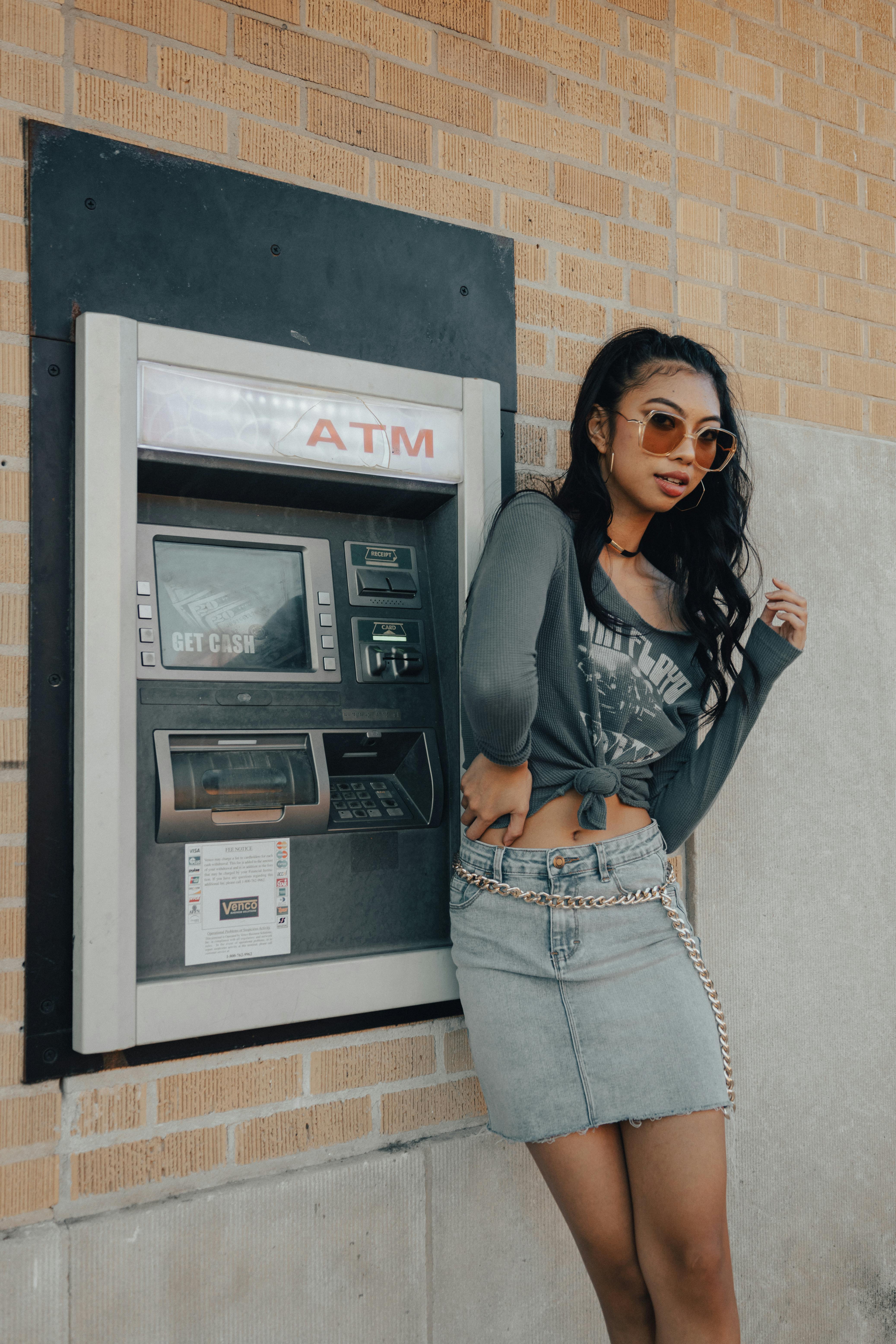 Fashionable young woman in sunglasses using an ATM, showcasing urban style.