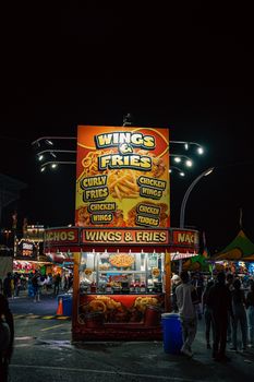 Vibrant food stall at a nighttime carnival offering wings and fries with illuminated signage.