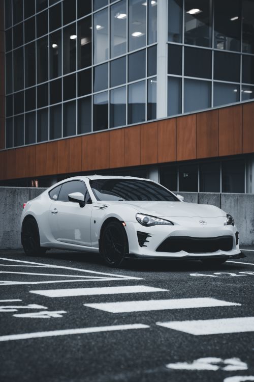 Free A white sports car is parked in a modern urban area with a tall office building backdrop. Stock Photo