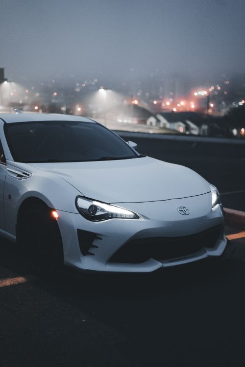 Free A white sports car with illuminated headlights parked at night in an urban area. Stock Photo