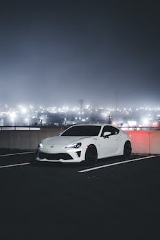 White car parked on a rooftop with city lights in the background, creating a moody nighttime atmosphere.