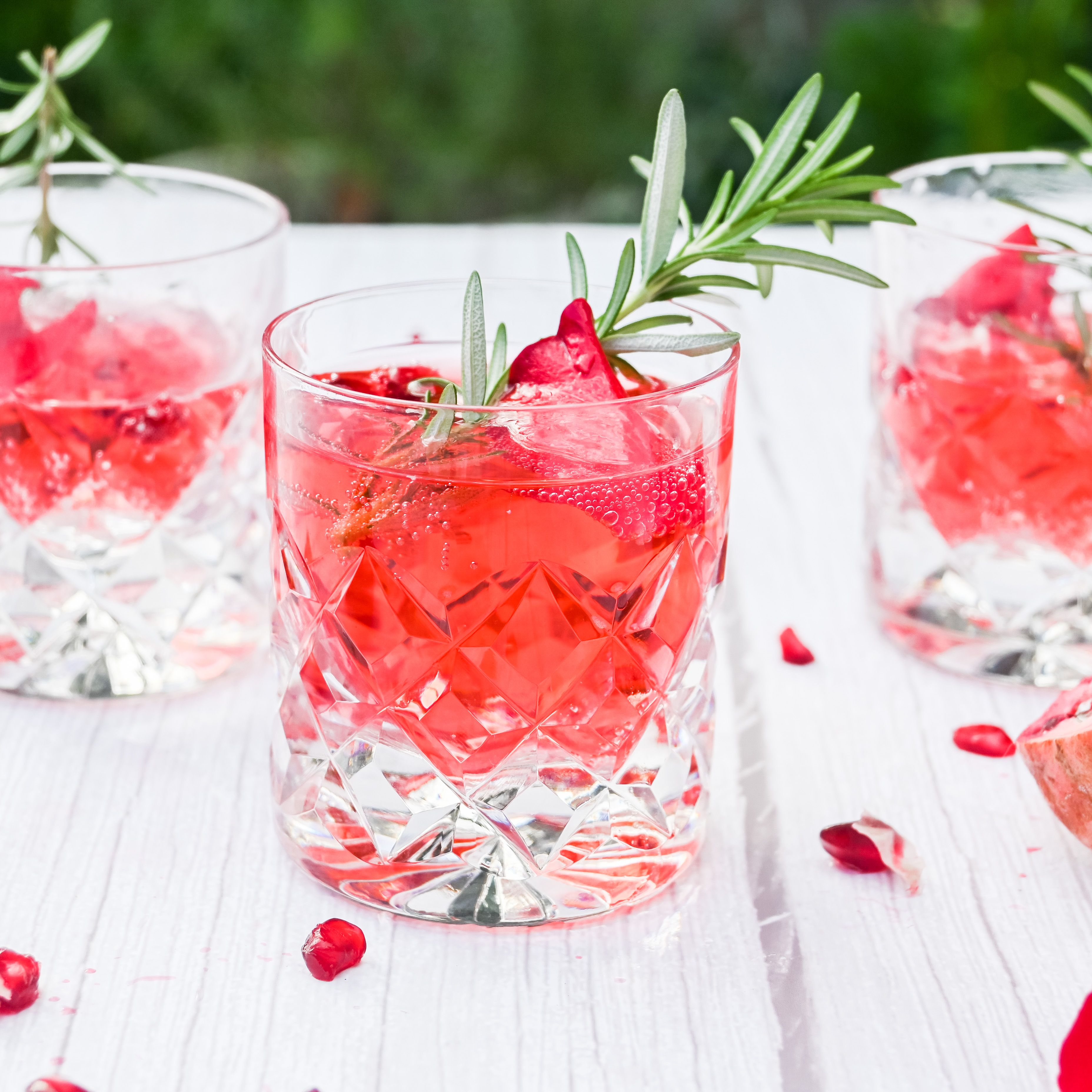 Crystal glasses of rose pomegranate cocktails garnished with rosemary and ice on a white table.
