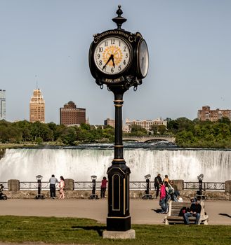 A vintage street clock stands prominently before the Niagara Falls as people enjoy the sunlit view in an urban setting.