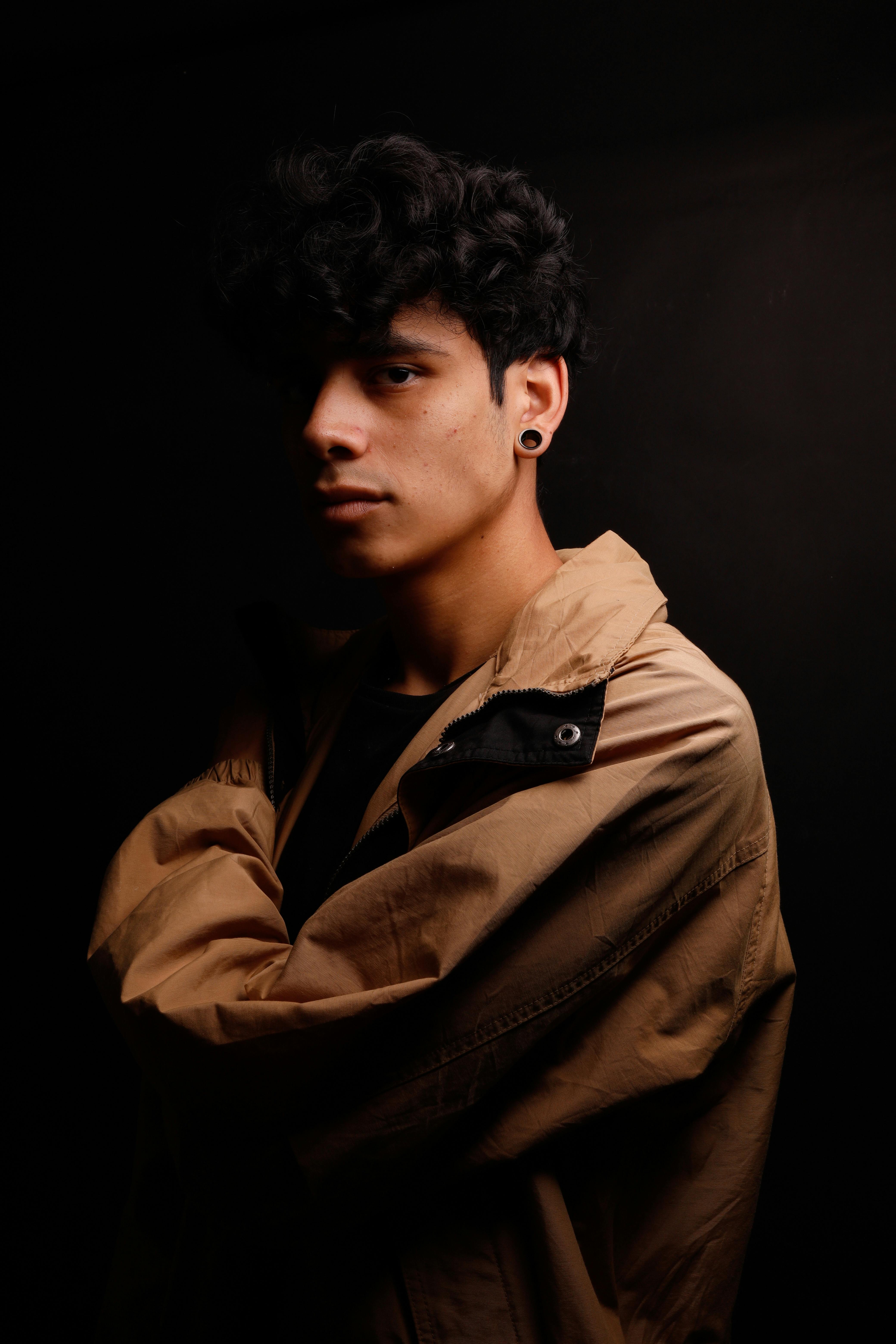 Moody studio portrait of a young man in a brown jacket with dark hair.