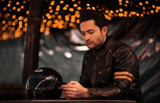 Young man in a leather jacket sits with his helmet in a dimly lit setting, reminiscent of a bar or club.