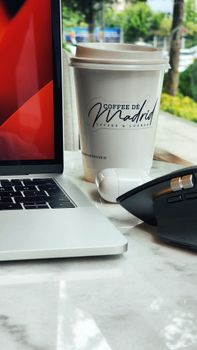 Close-up of coffee cup with laptop and peripherals on a marble desk, showcasing a workspace setup.