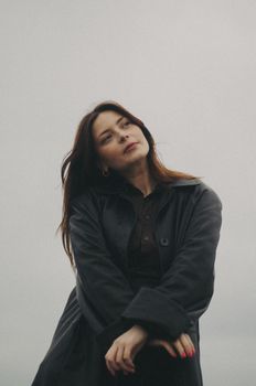 A thoughtful woman poses elegantly in a black coat against a neutral background.