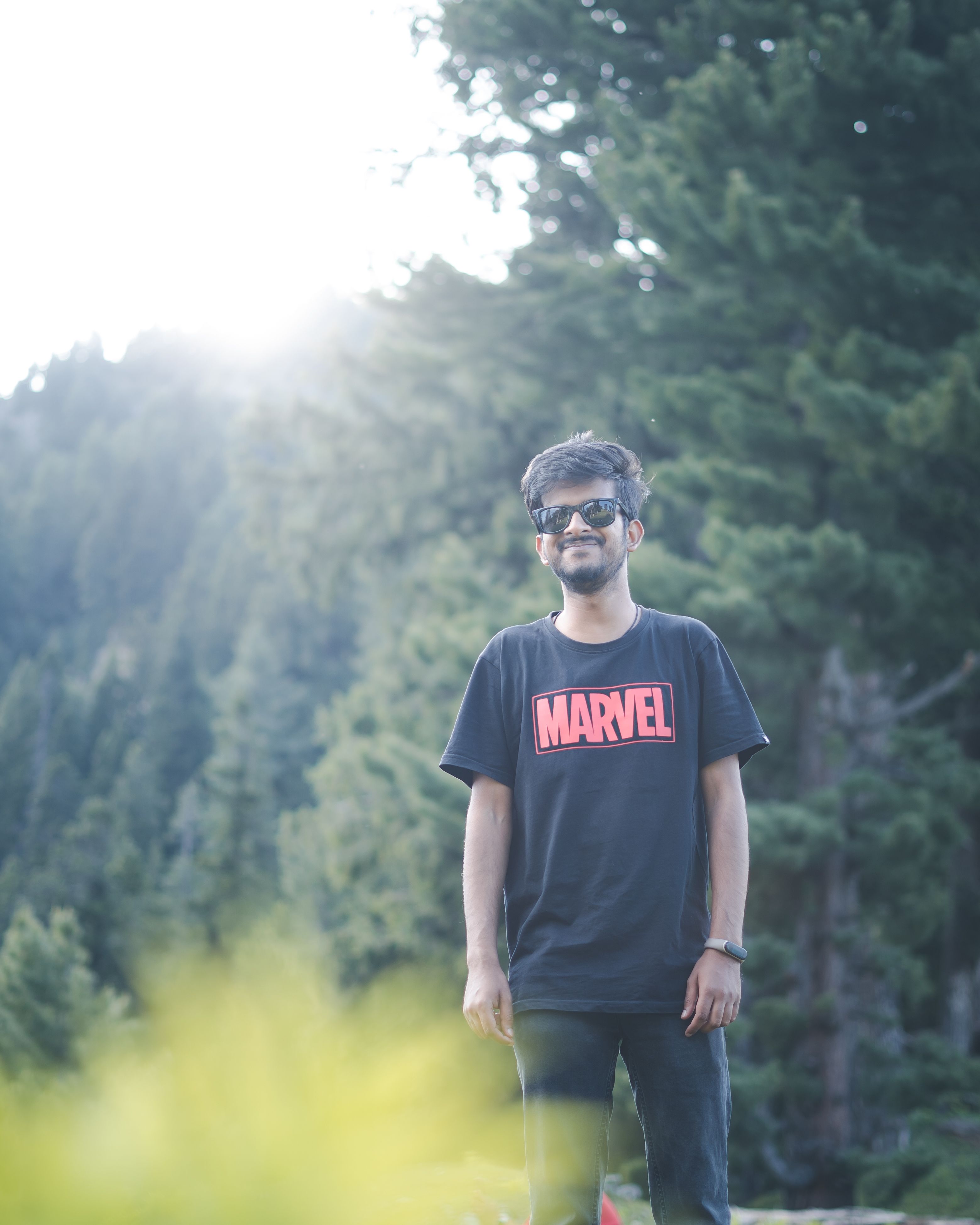 A smiling young man wearing sunglasses stands in a lush forest in Srinagar.