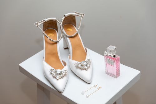 Free White bridal shoes adorned with crystals alongside pink perfume bottle and pearl earrings on a table. Stock Photo