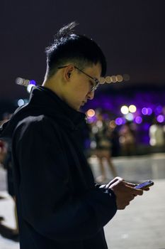 A young man illuminated by streetlights, texting on his smartphone in an urban setting at night.