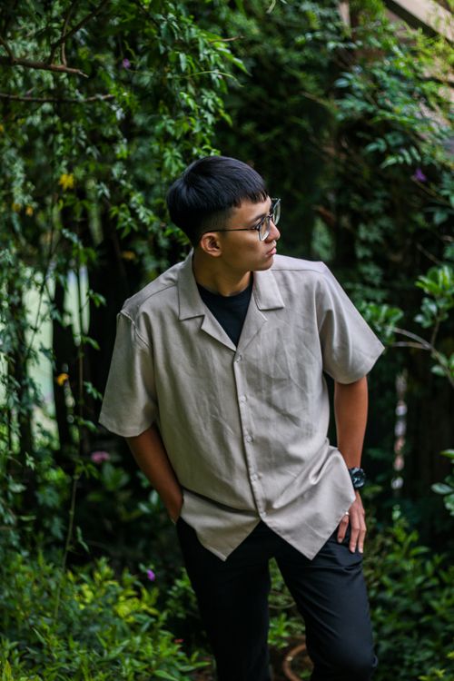 Free Portrait of a young man standing among dense greenery, wearing eyeglasses and a casual shirt. Stock Photo