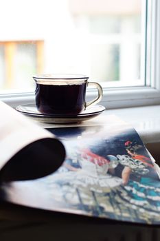 A close-up of a coffee cup on a windowsill next to an open magazine.