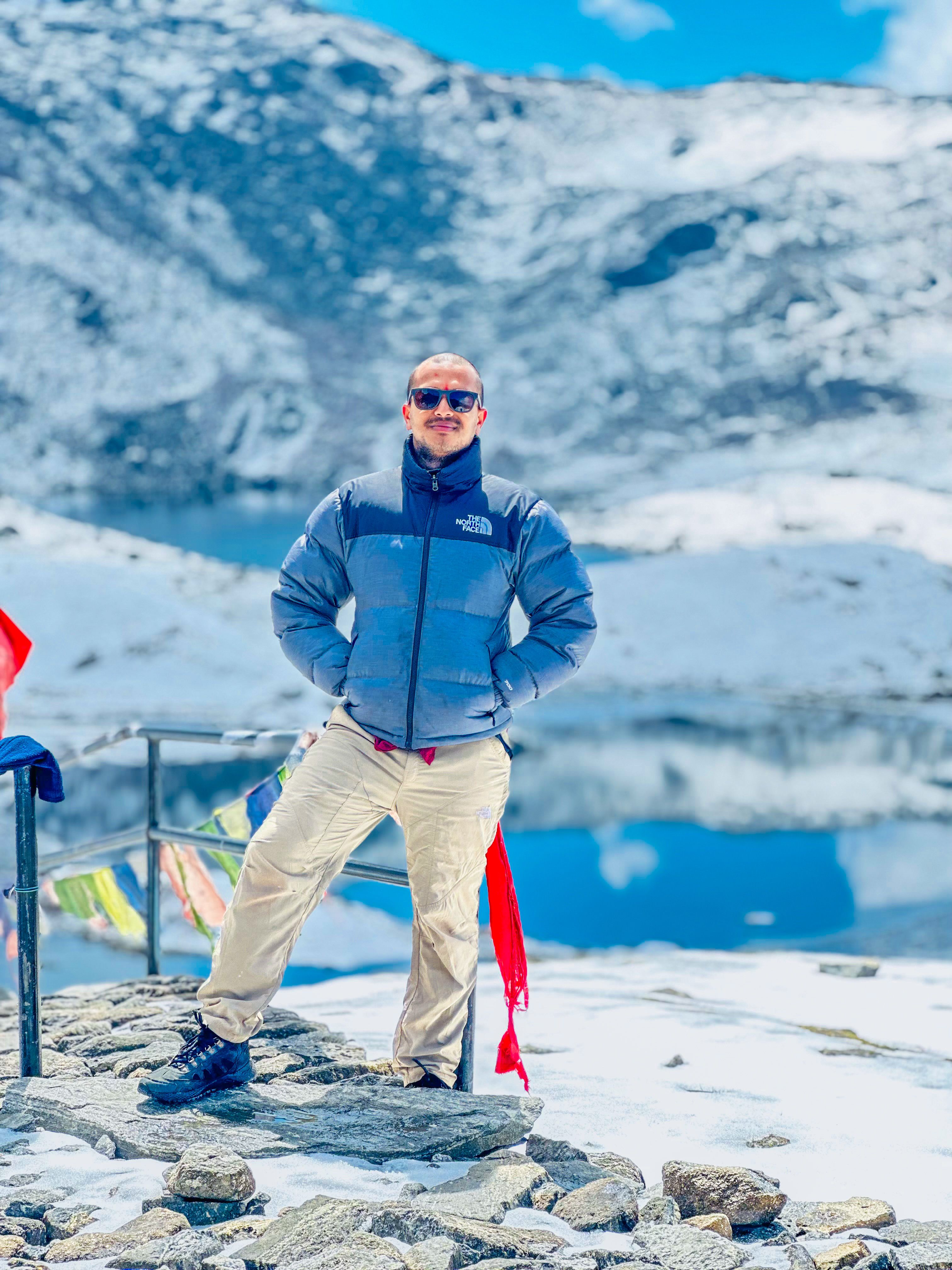 Man in winter gear posing on snowy mountain path with scenic lake and mountains in background.