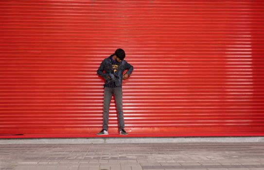 Man with short hair in denim jacket stands against a red urban wall, looking down. Street style and modern vibe.