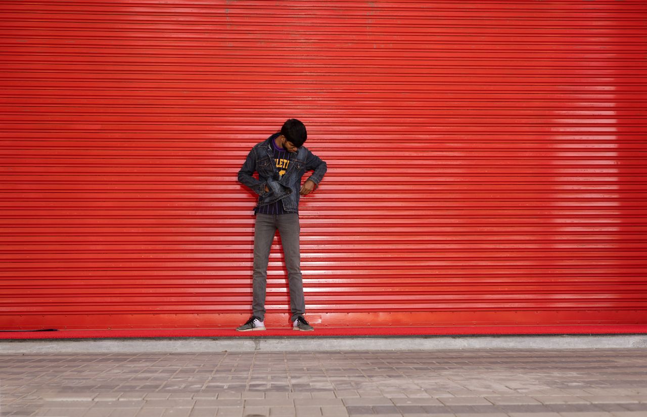 man with short hair in denim jacket stands against a red urban wall lo