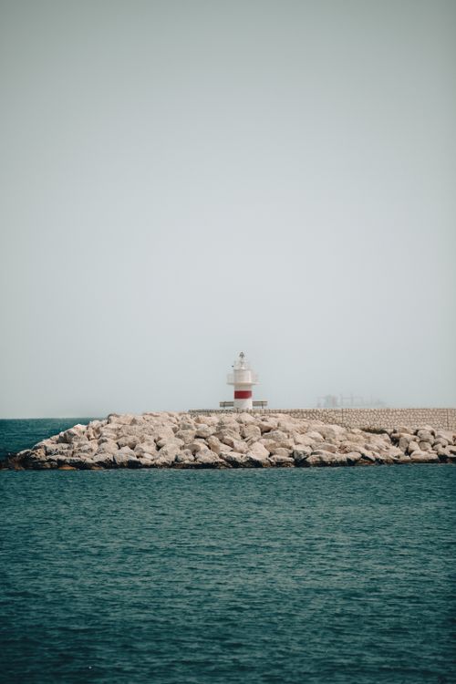 Free Peaceful view of a lighthouse on a rocky shore under a clear summer sky. Stock Photo
