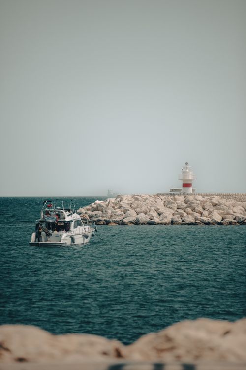 Free A serene ocean scene featuring a lighthouse, motorboat, and rocky shore. Stock Photo