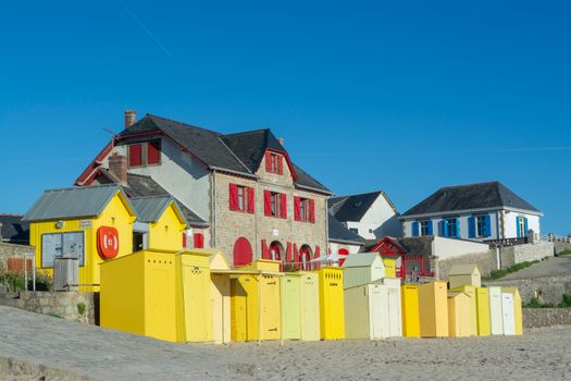Vibrant beach huts line the sandy shore in Batz-sur-Mer, France, under a clear blue sky.