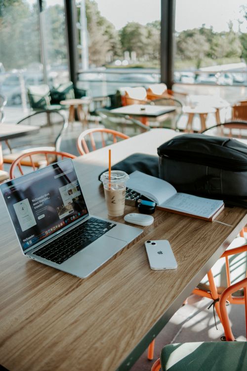 Free Cozy workspace in a café featuring a laptop, smartphone, and iced coffee on a sunny day. Stock Photo