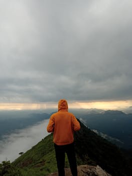Solitary hiker in orange hoodie stands atop mountain ridge under dramatic sky.