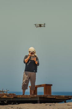 A person controlling a drone on a sandy beach with clear skies and wooden benches.