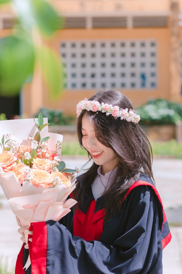 Girl In Graduation Gown And Flowers Wreath Holding Bouquet