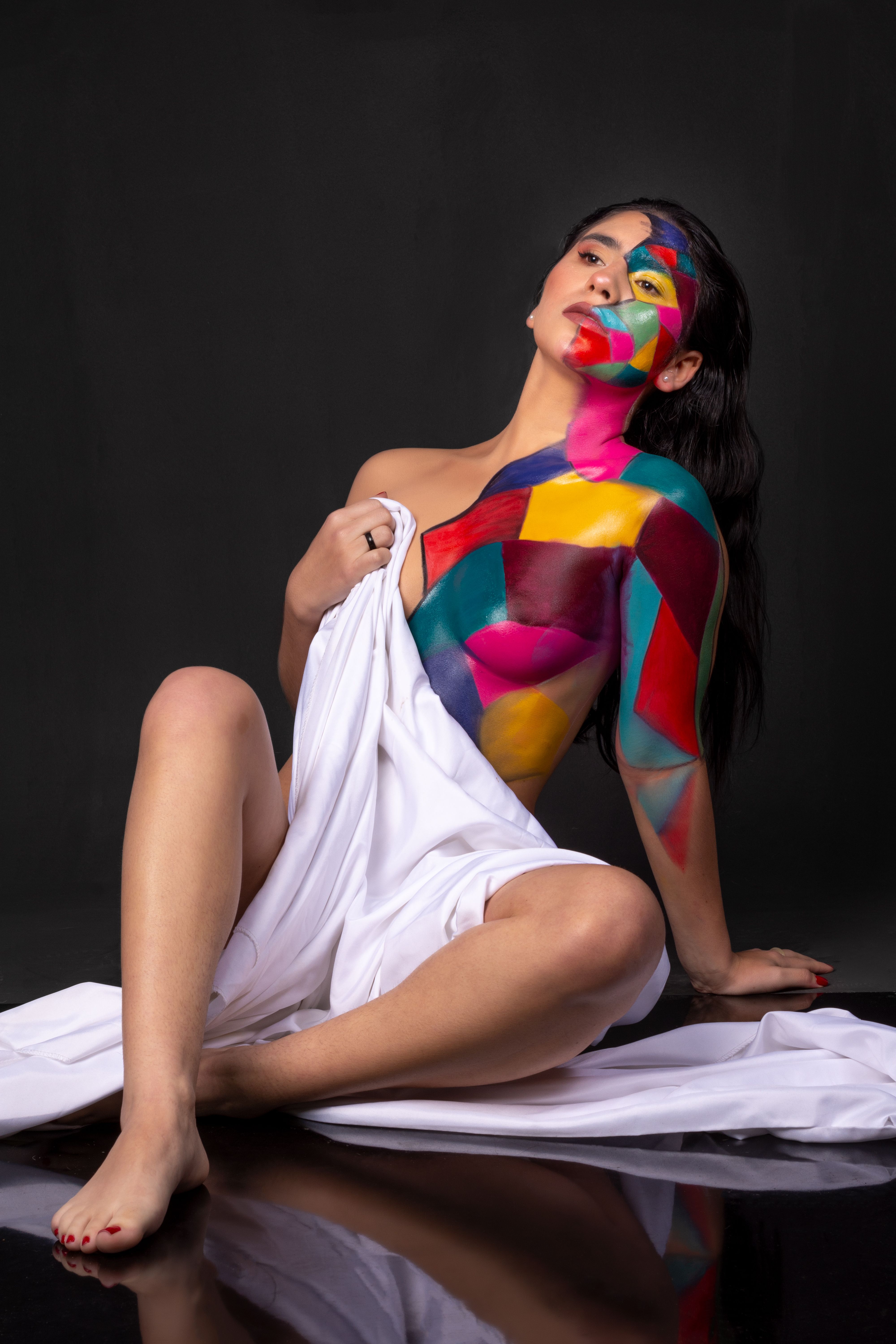 Creative studio portrait of a woman with colorful geometric body painting sitting on black background.