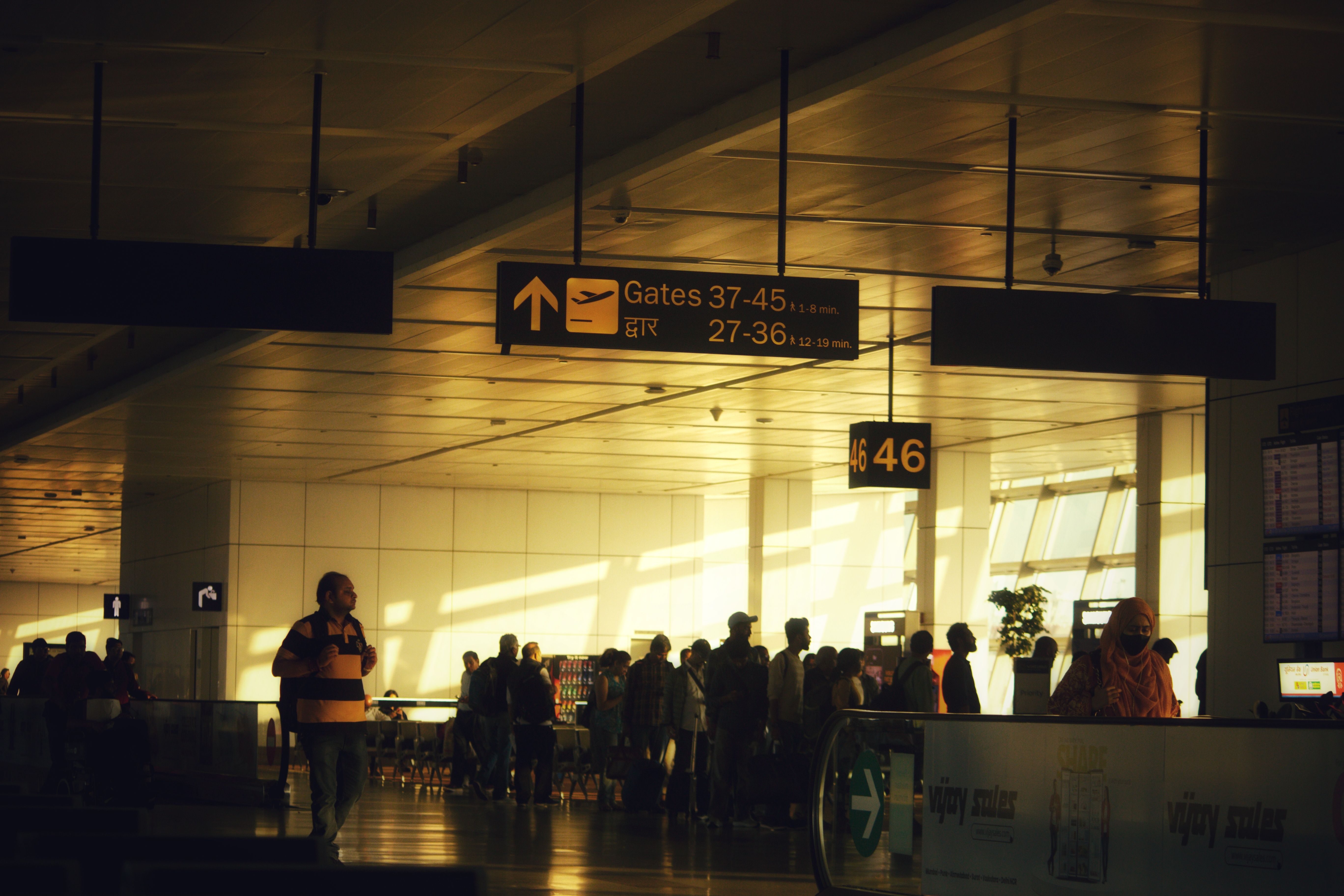 Passengers wait beneath airport gate signs in a terminal filled with warm evening light