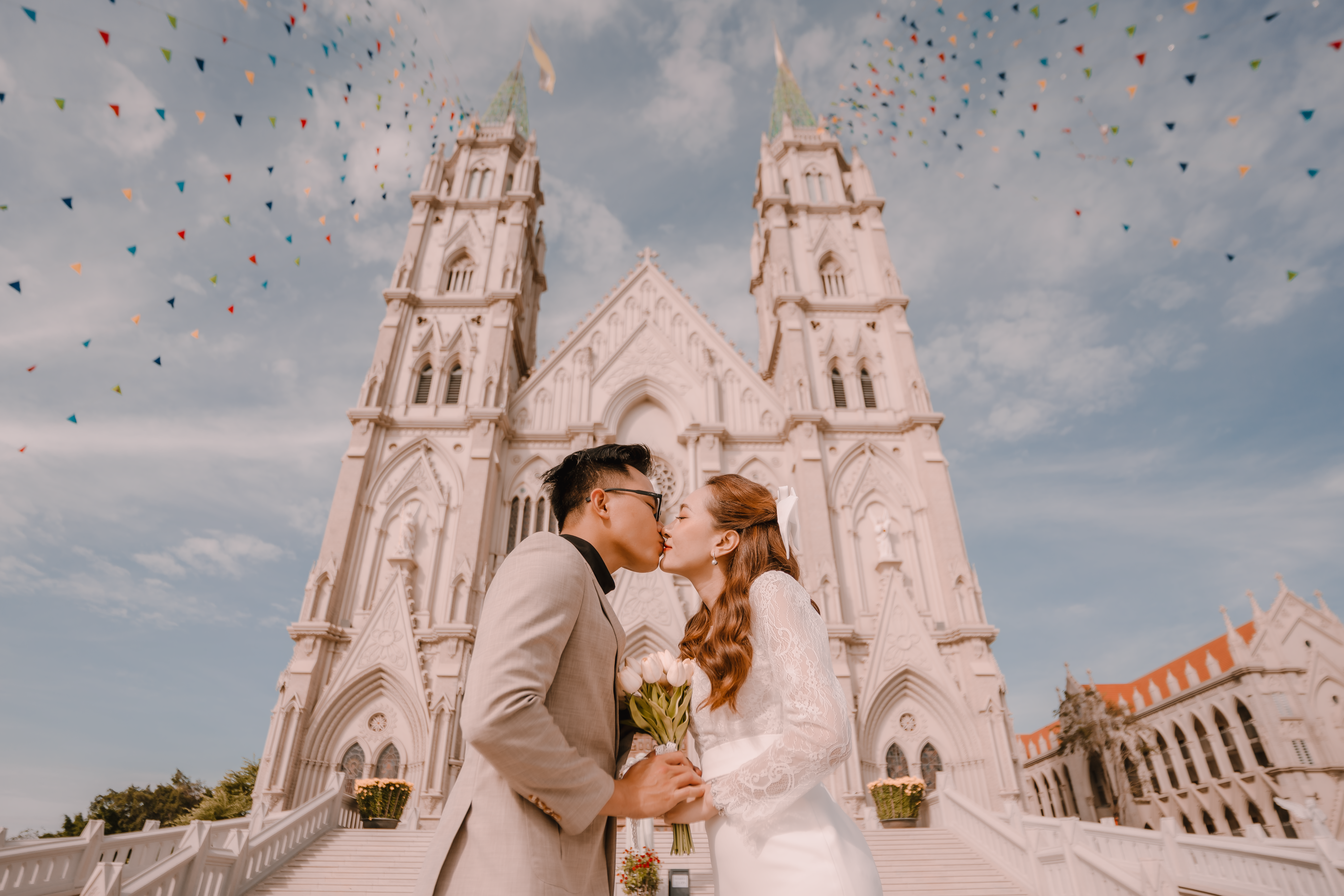 A newlywed couple kisses in front of a grand Gothic cathedral under a colorful sky.