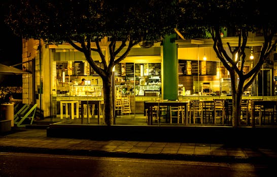 Exterior of a warmly lit urban café at night, featuring empty outdoor seating and trees lining the pavement.